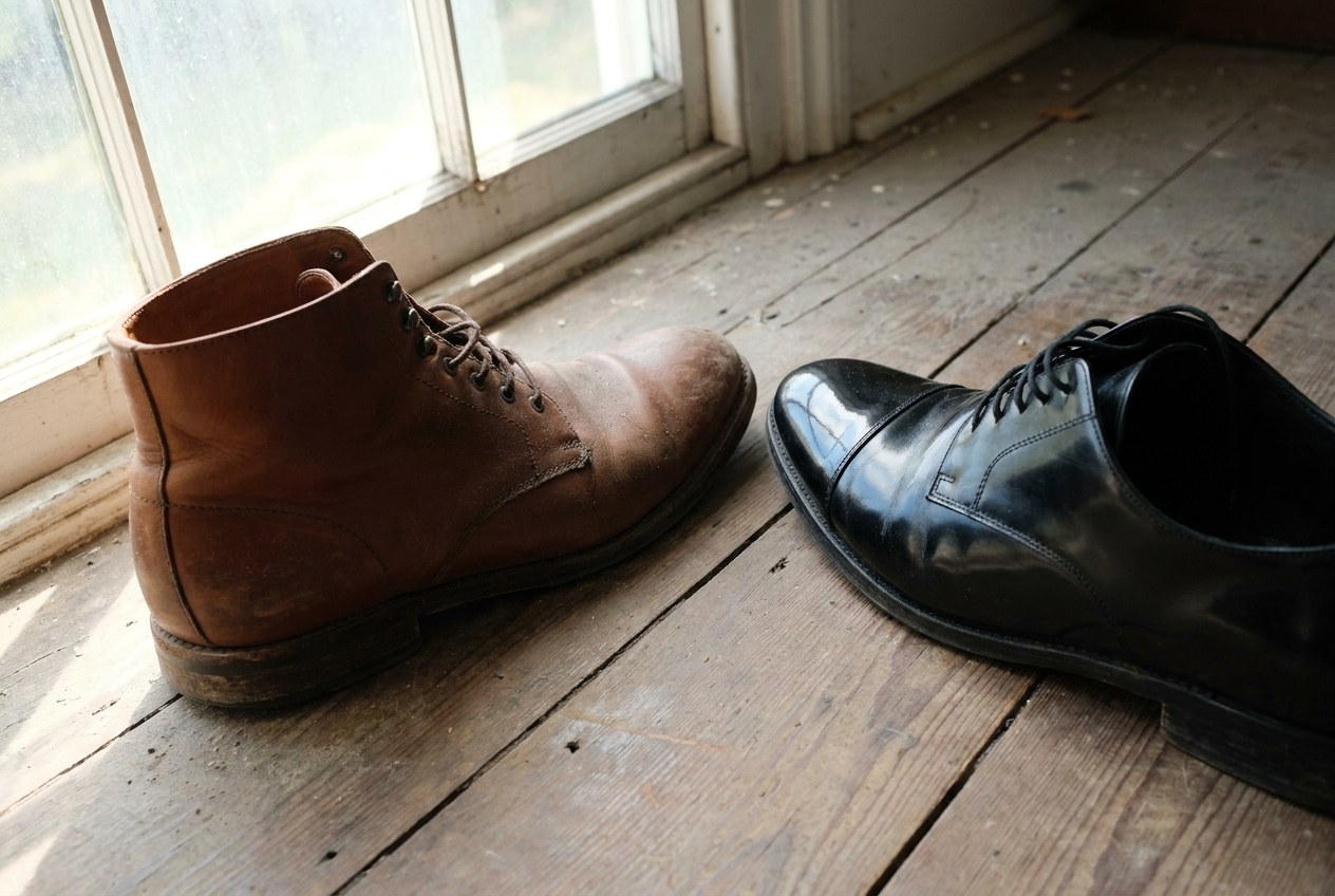 Zwei Lederschuhe nebeneinander im Seitenlicht, links eine matte braune Oberfläche ohne Reflex, rechts eine schwarze Kappe, die das Fenster scharf spiegelt
