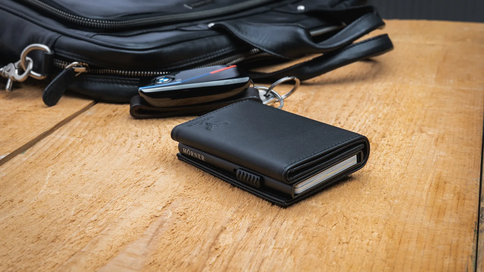Schwarzes Hörner Nexus Slim Wallet auf einem Holztisch neben einer Ledertasche und einem Autoschlüssel, ein Pop-Up-Kartenwallet mit Easy-Slide