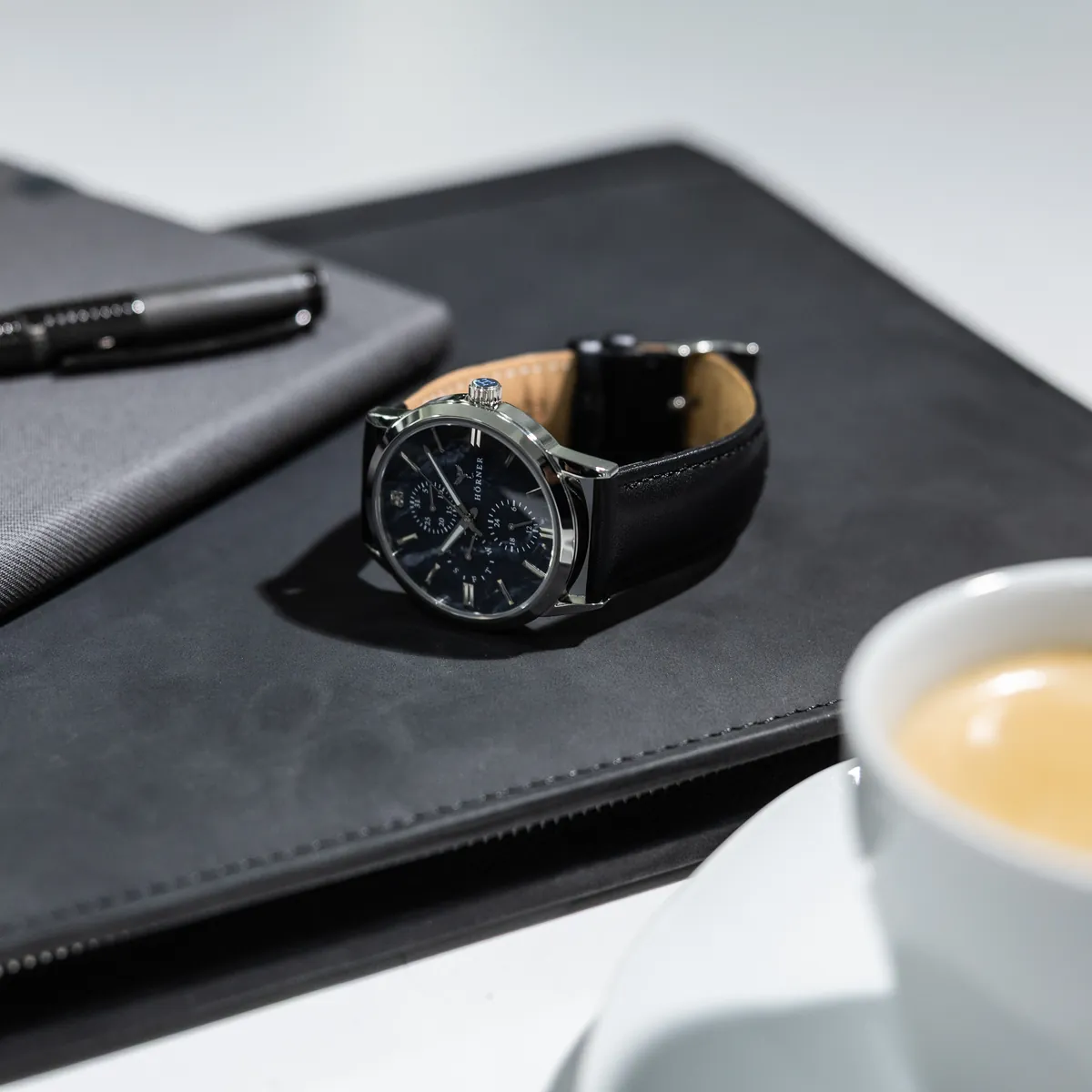 Hörner Nova Midnight Quarzuhr mit dunkelblauem Zifferblatt in Perlmutt-Optik und schwarzem Lederband auf einer dunklen Schreibmappe, daneben ein Stift und eine Kaffeetasse