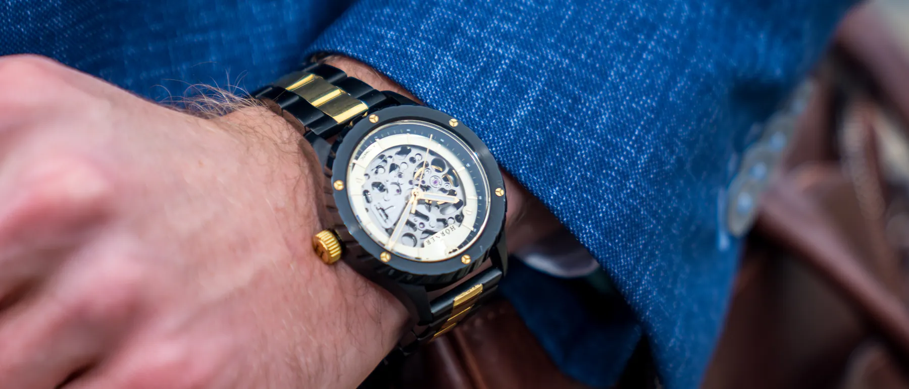Hörner Pulsar Imperial Automatikuhr mit schwarzem Gehäuse und goldenen Akzenten am Handgelenk, im Hintergrund eine braune Ledertasche