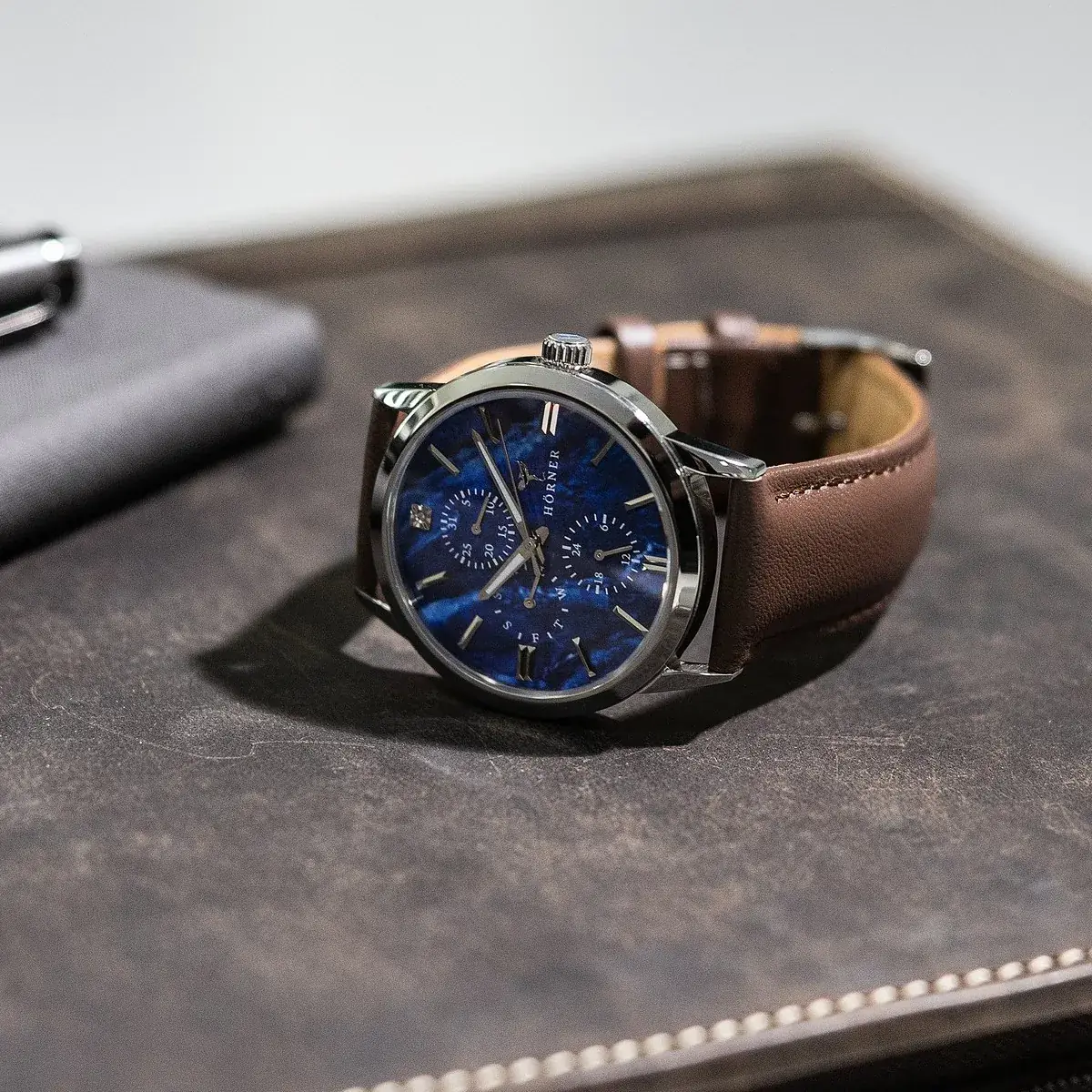 Hörner Nova Nebula Quarzuhr mit Zifferblatt in blauer Perlmutt-Optik, Hilfszifferblättern und braunem Lederarmband auf einer dunklen Ledermappe