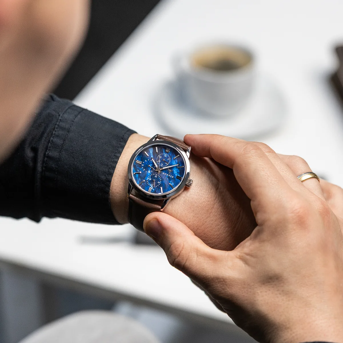 Hörner Nova Nebula mit blauem Zifferblatt und braunem Lederband am linken Handgelenk, die rechte Hand mit Ehering greift an die Krone bei 3 Uhr, im Hintergrund unscharf eine Kaffeetasse auf einem Tisch